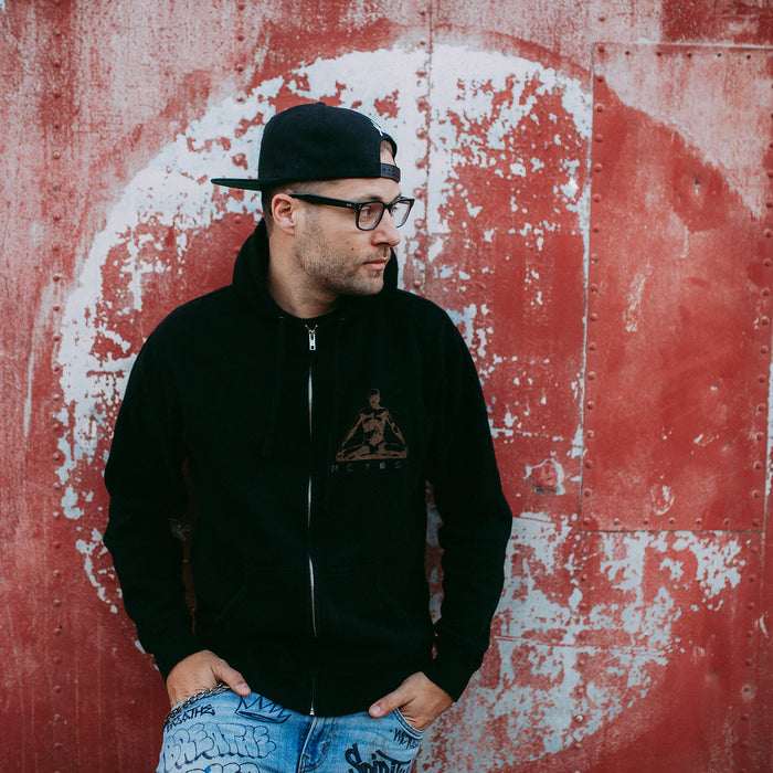 A man in an MC YOGI black hoodie, glasses, and a backwards cap stands before a red wall with peeling white paint, looking to the side with one hand in his pocket.