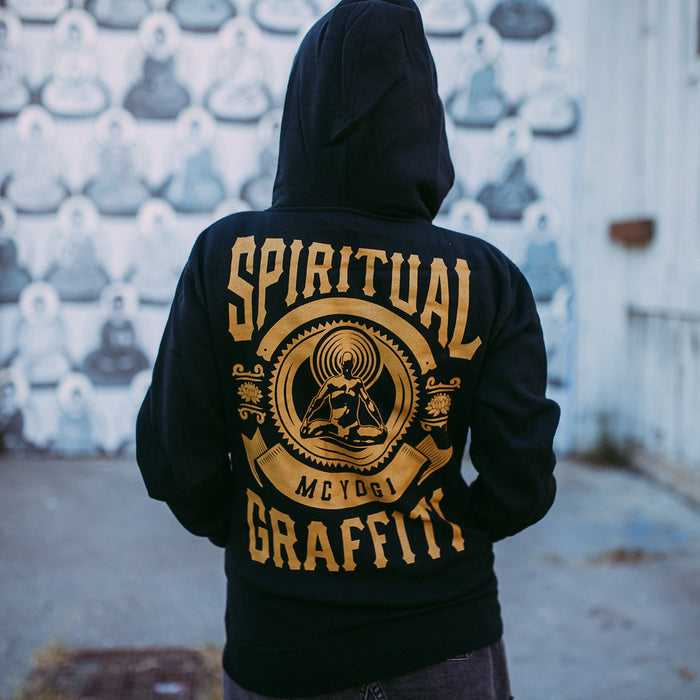 A person wearing an MC YOGI black hoodie with gold SPIRITUAL GRAFFITI lettering and a graphic design stands in front of a wall covered in monochrome portraits.